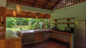 A kitchen with wooden cabinets, a sink, a stove, and a refrigerator. There is a window overlooking a garden. - Tucan Chiquita Beach Casitas (Playa Chiquita)