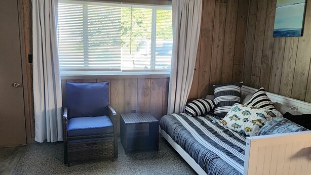 Ein gemütliches Zimmer mit einer Holzvertäfelung, einem Fenster mit Vorhängen, einem gestreiften Sofa mit Kissen und einem Korbstuhl.