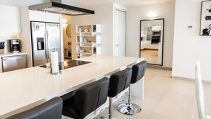 Cozinha moderna com ilha central, banquetas, eletrodomésticos de aço inoxidável e painel de fundo branco.
