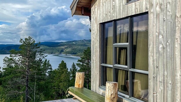 A wooden cabin with large windows overlooking a lake and forested hills.