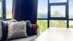 A room with a sofa, a table, and a view of green hills through large windows.