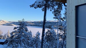 A snowy landscape with a lake and mountains in the background, viewed from a wooden cabin.