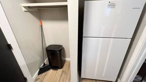 A compact kitchen with a white refrigerator, a broom, and a trash bin. - Cozy 1 Bedroom Rental With Den Free Parking Close to University (Edmonton)