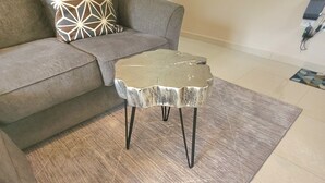 Ein Wohnzimmer mit grauem Sofa, einem gemusterten Zierkissen, einem kleinen metallischen Tisch mit schwarzen Beinen und einem grauen Teppich mit Muster.