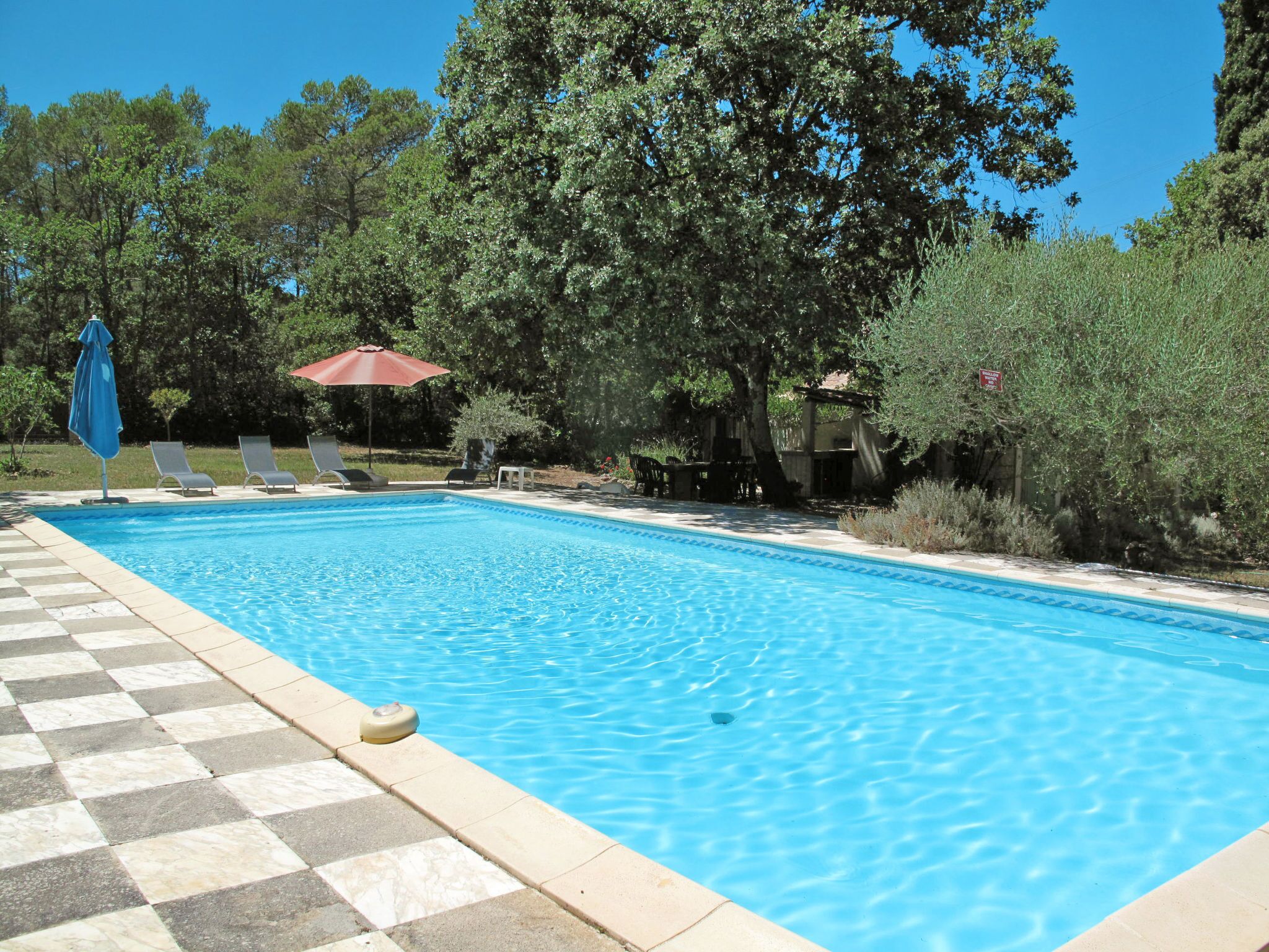 A rectangular swimming pool with a checkered tile border, surrounded by lounge chairs and a red umbrella.