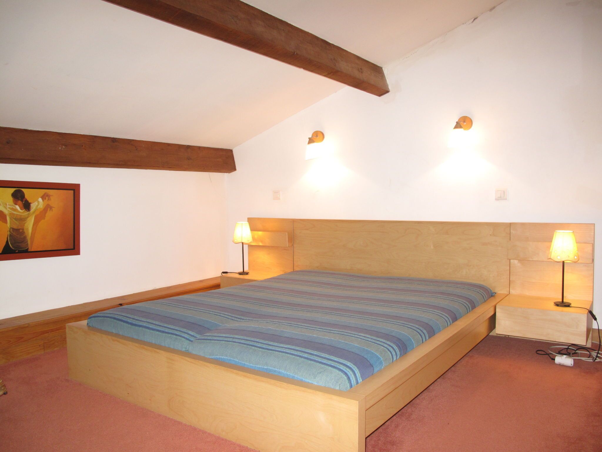 Ein Schlafzimmer mit hölzernem Bettgestell, geneigter Decke mit freiliegenden Balken, zwei Nachttischlampen und einem Bild an der Wand.
