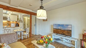 A cozy living room with a wooden floor, a sofa, a coffee table with a vase of flowers, a TV, and a kitchen area visible through an open door.