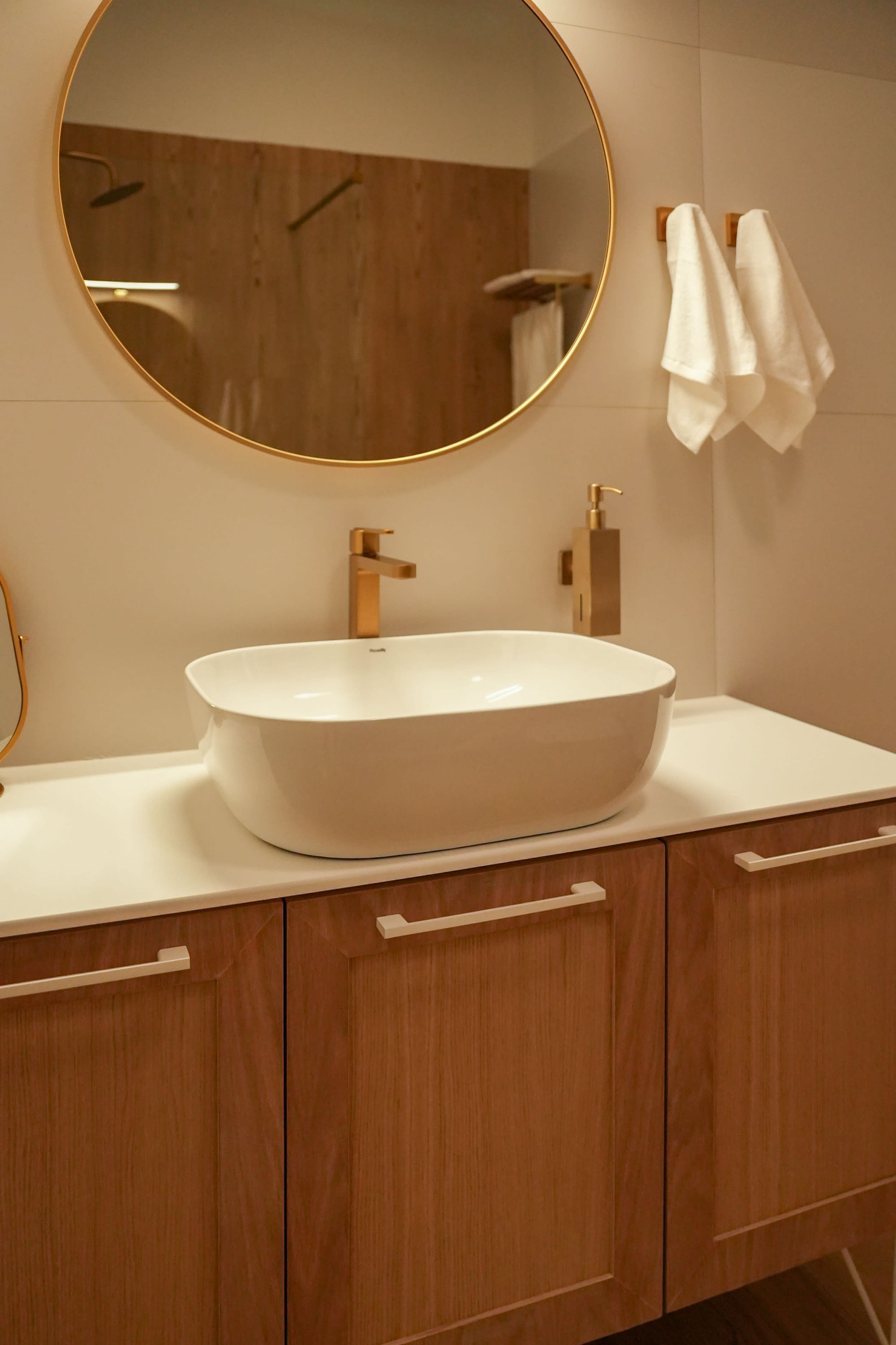 Een badkamer met een ronde spiegel, een witte wastafel, houten kastjes en een handdoekhouder.