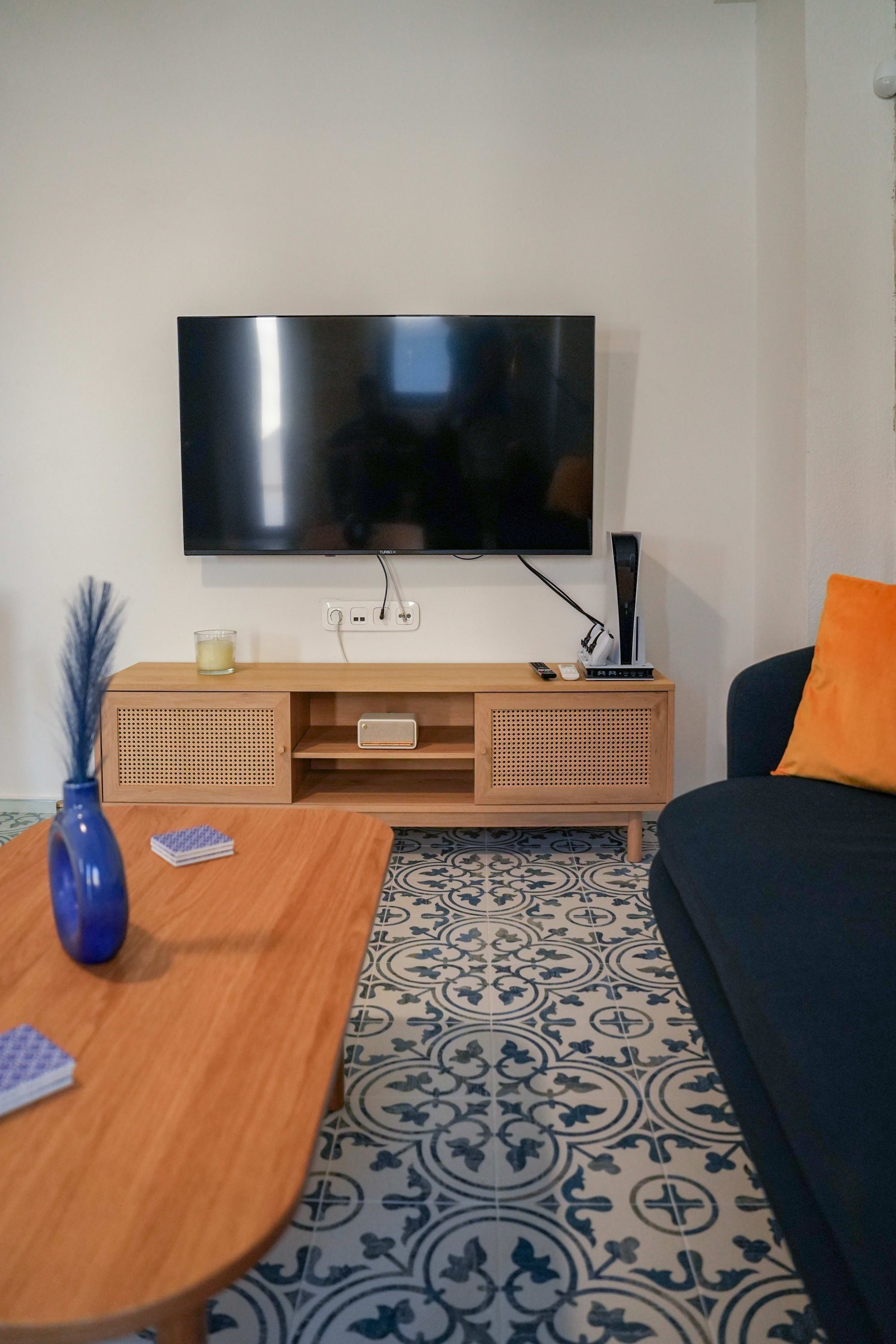 A living room with a flat-screen TV mounted on the wall, a wooden entertainment unit below, a patterned rug, a blue vase, and a dark-colored sofa with an orange pillow.