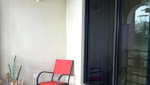 Un balcón con una mesa redonda, una silla roja, una planta en maceta y un ventanal grande.