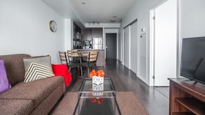 Un salon moderne avec un canapé, une table basse et un meuble pour la télévision. On y trouve également un coin repas avec une table et des chaises, et une cuisine à l’arrière-plan.