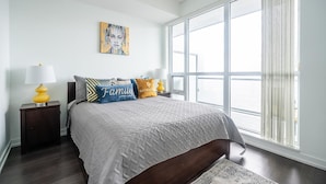 Une chambre avec une grande fenêtre, un lit recouvert d’une couette grise et agrémenté de coussins décoratifs.