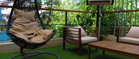 A hanging chair, a wooden sofa, and a table on a balcony with artificial grass and a view of lush greenery.