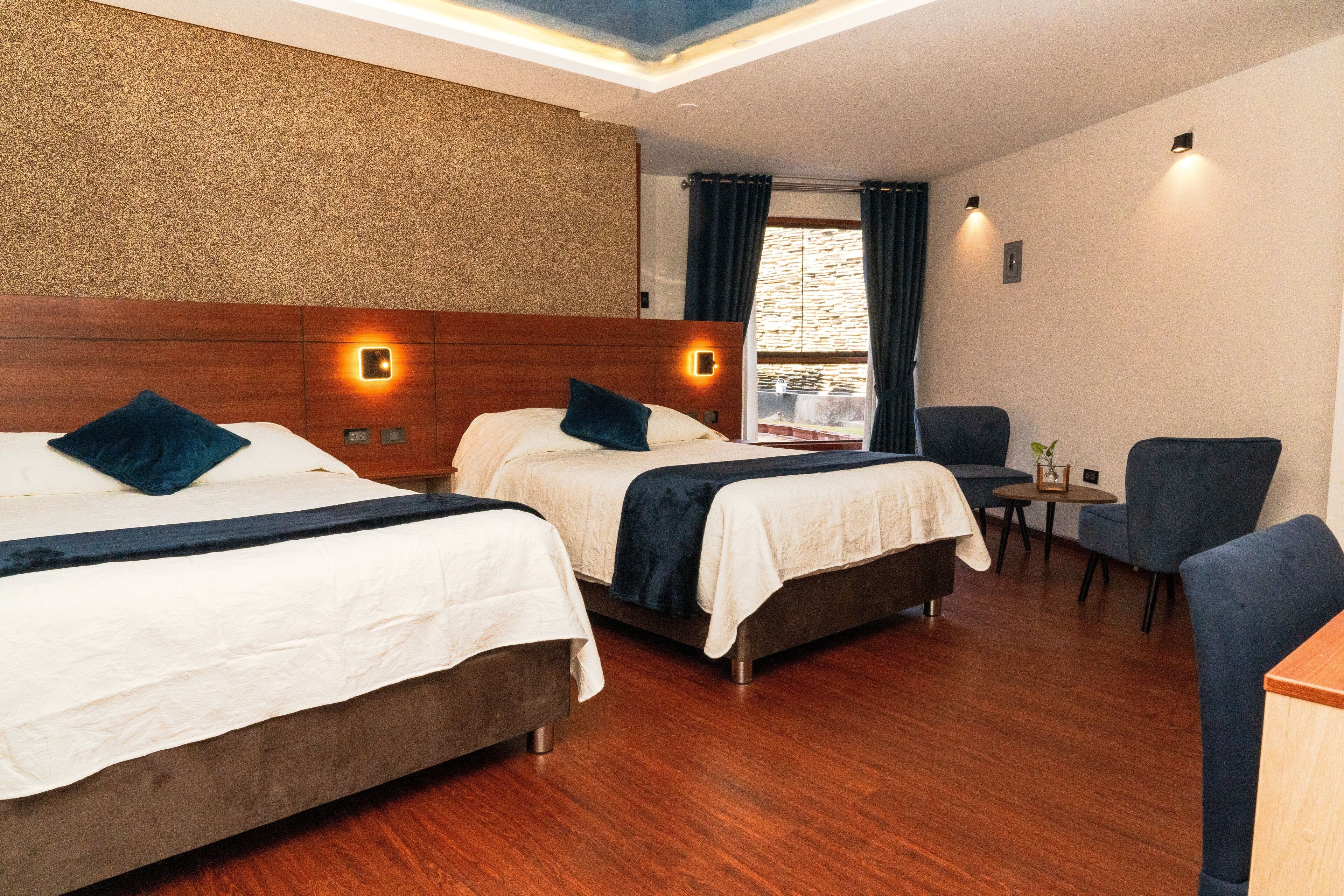 A hotel room with two beds, a small table, a chair, and a window with curtains.