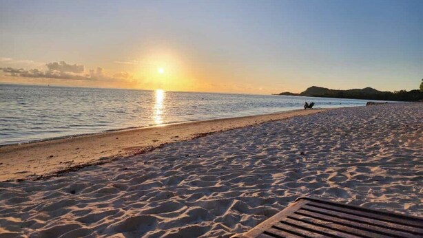 A beach with a wooden bench in the foreground and a sunset in the background.