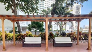 A covered outdoor seating area with two cushioned benches, surrounded by potted plants and a tiled floor.