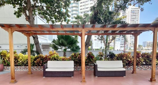 A covered outdoor seating area with two cushioned benches, surrounded by potted plants and a tiled floor.