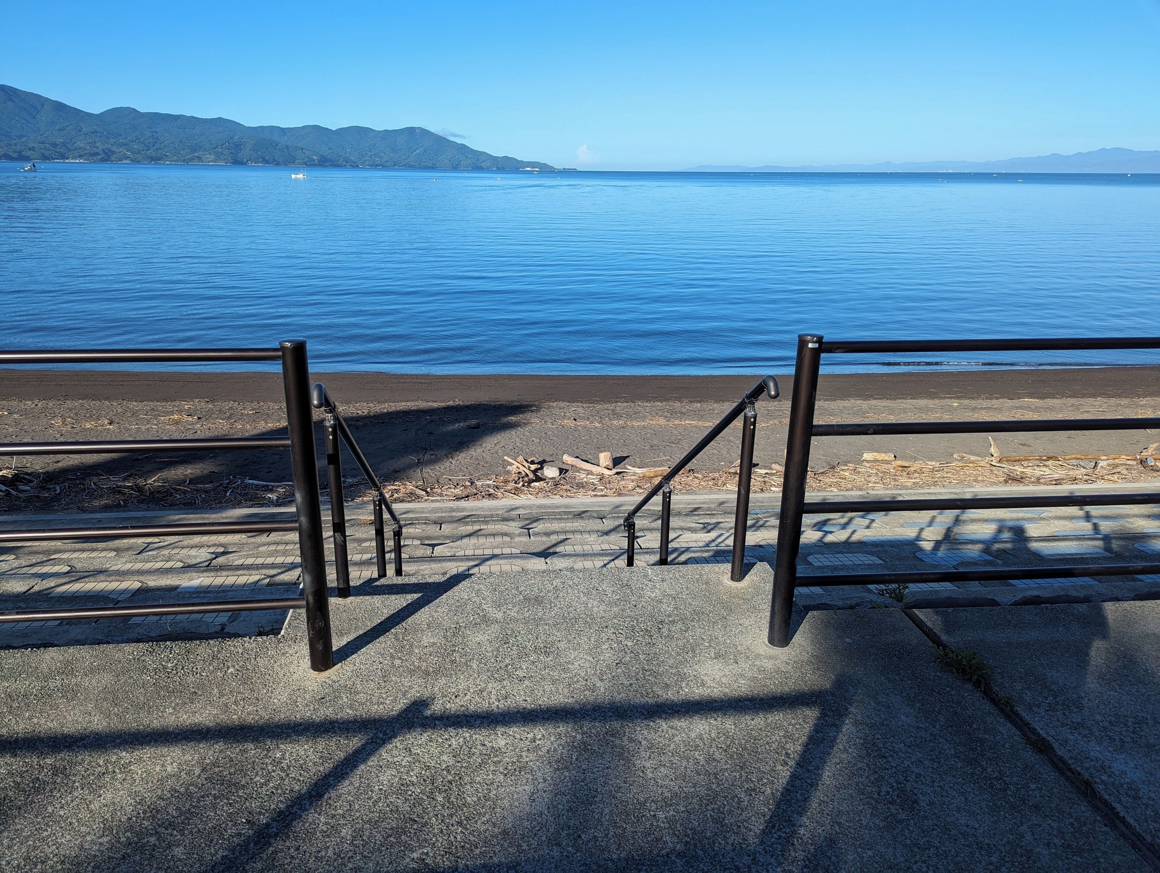 A concrete staircase leading to a beach with a metal railing, overlooking a calm body of water and distant mountains.