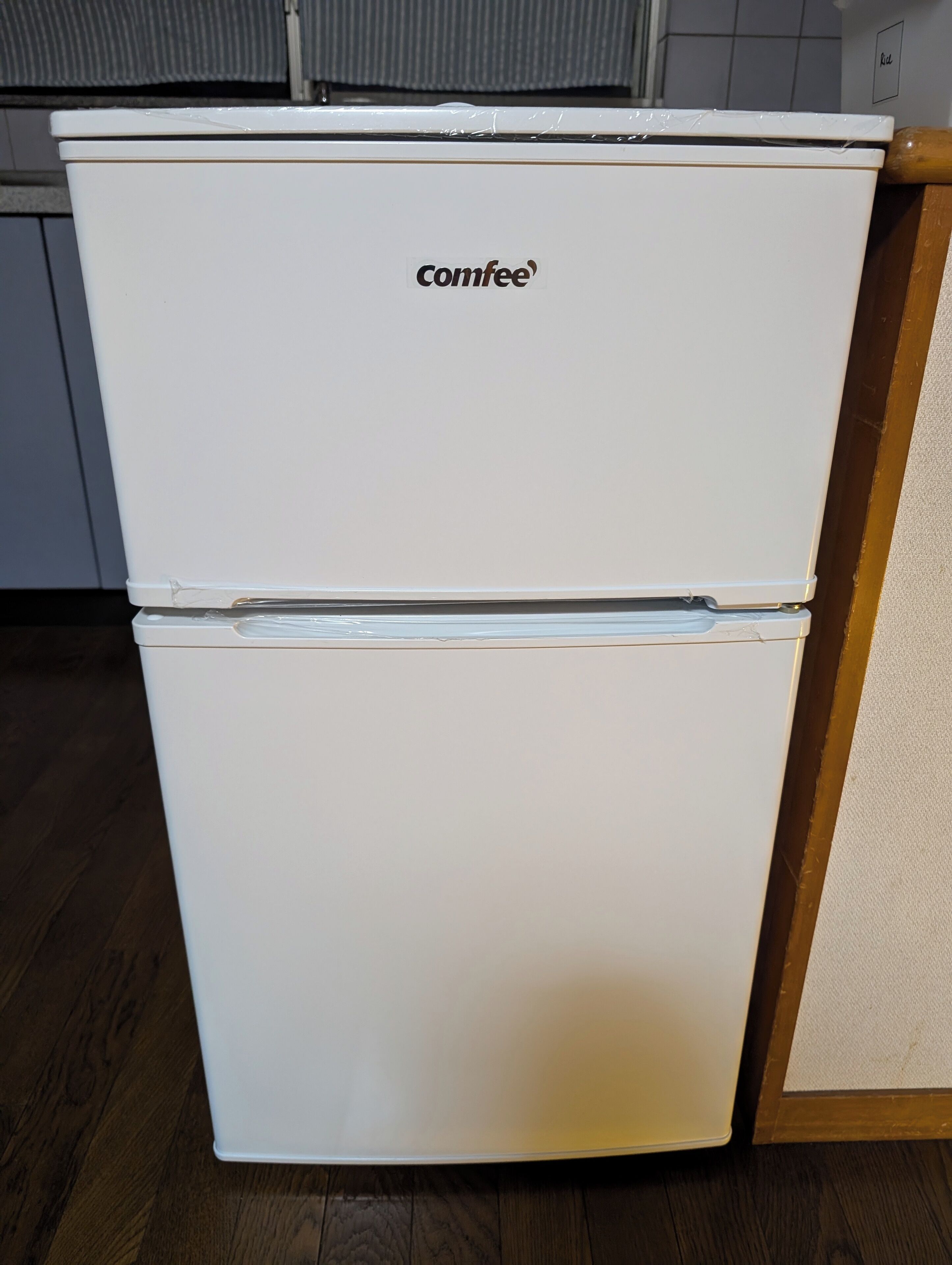 A white Comfee refrigerator in a kitchen with wooden flooring and a tiled backsplash.