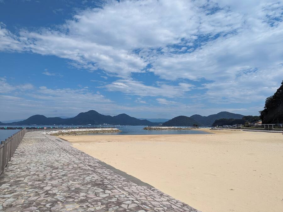 A beach with a stone walkway, calm water, and mountains in the background.