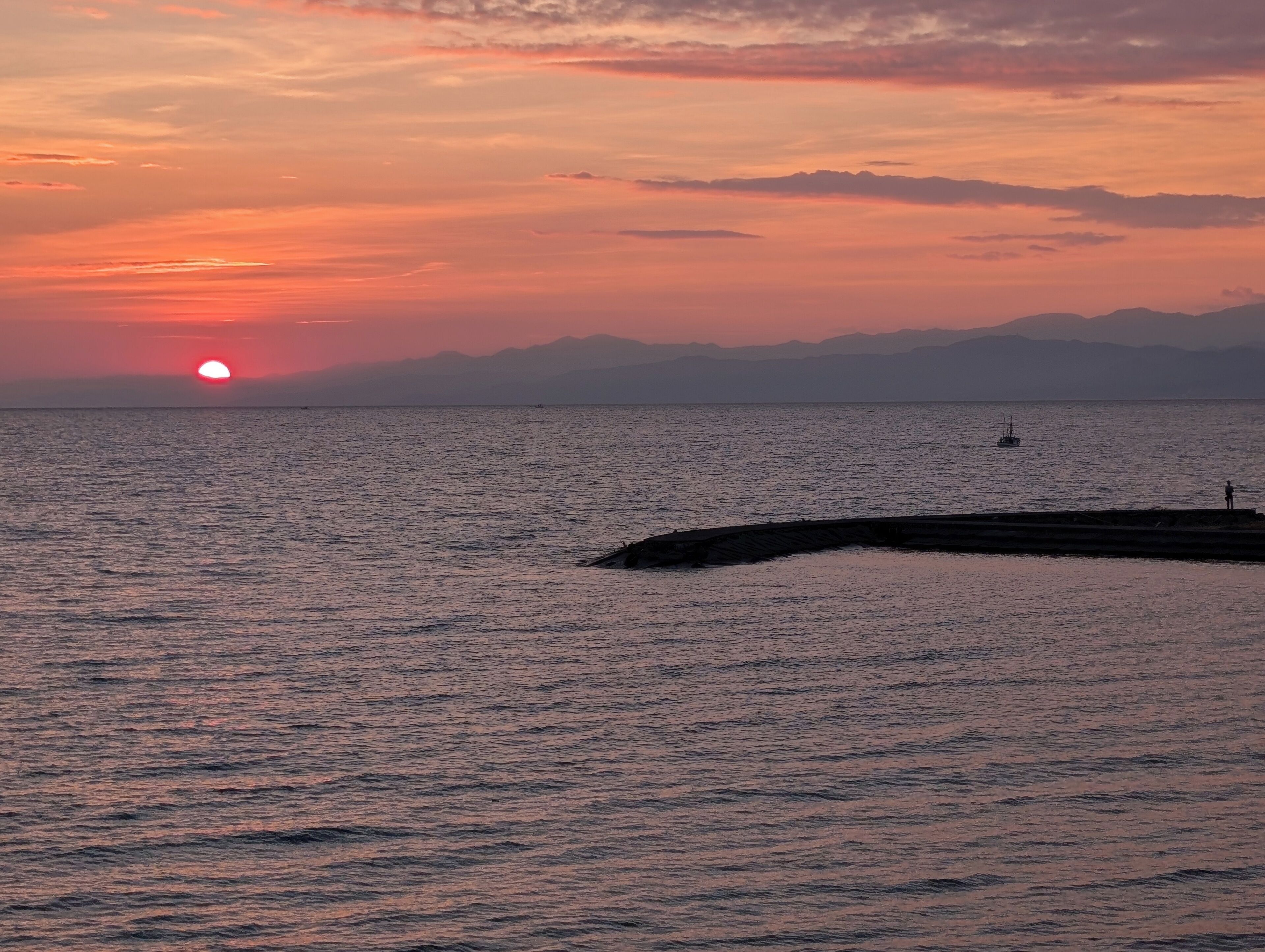 Un coucher de soleil sur une mer calme, avec une jetée qui s’avance dans l’eau.