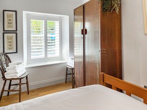 A bedroom with a bed, a wooden wardrobe, a window with blinds, and a chair with towels.