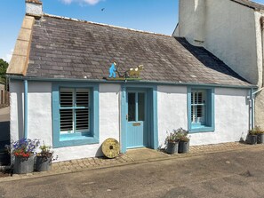 A cottage with a blue door, two windows with shutters, a small porch, and potted plants.