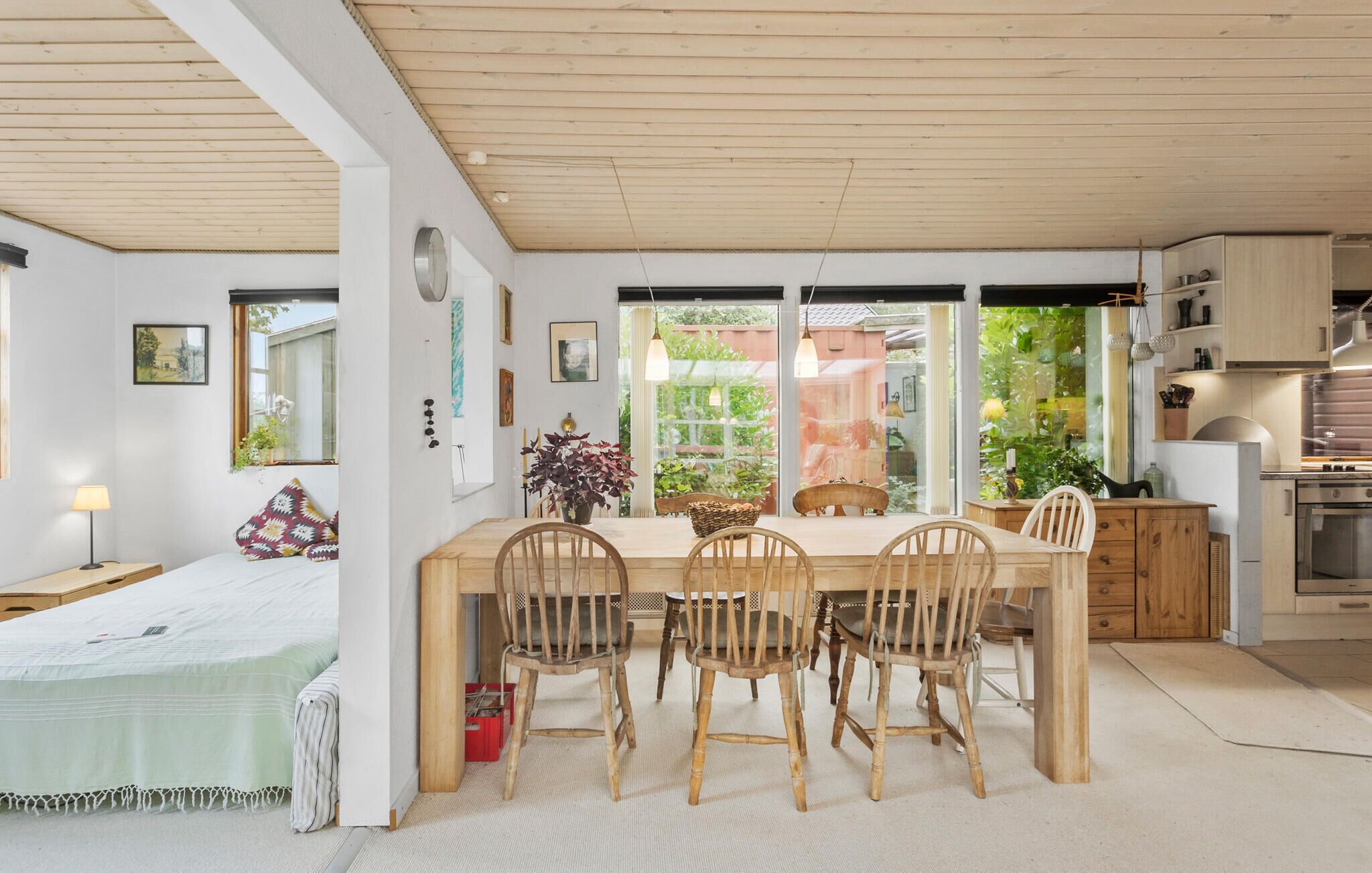 Une pièce chaleureuse et bien éclairée, avec une table à manger en bois, quatre chaises et un lit recouvert d’une couverture verte. Une fenêtre offre une vue sur la verdure environnante.