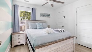 Una camera da letto con un letto, un comodino, una ventola a soffitto, una finestra con tende e una porta.