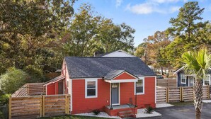A small, single-story red house with a dark roof, a front porch, and a wooden fence. - Entire Home - Great Location (Charleston)