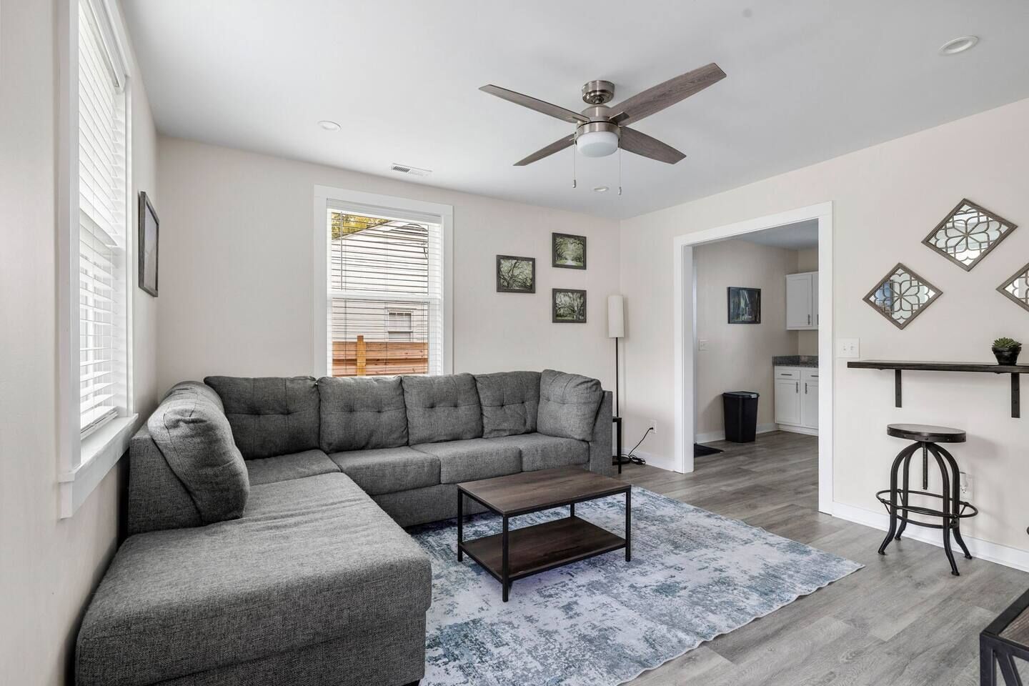 A modern living room with a grey sectional sofa, a coffee table, and a ceiling fan. There is a kitchen visible in the background.