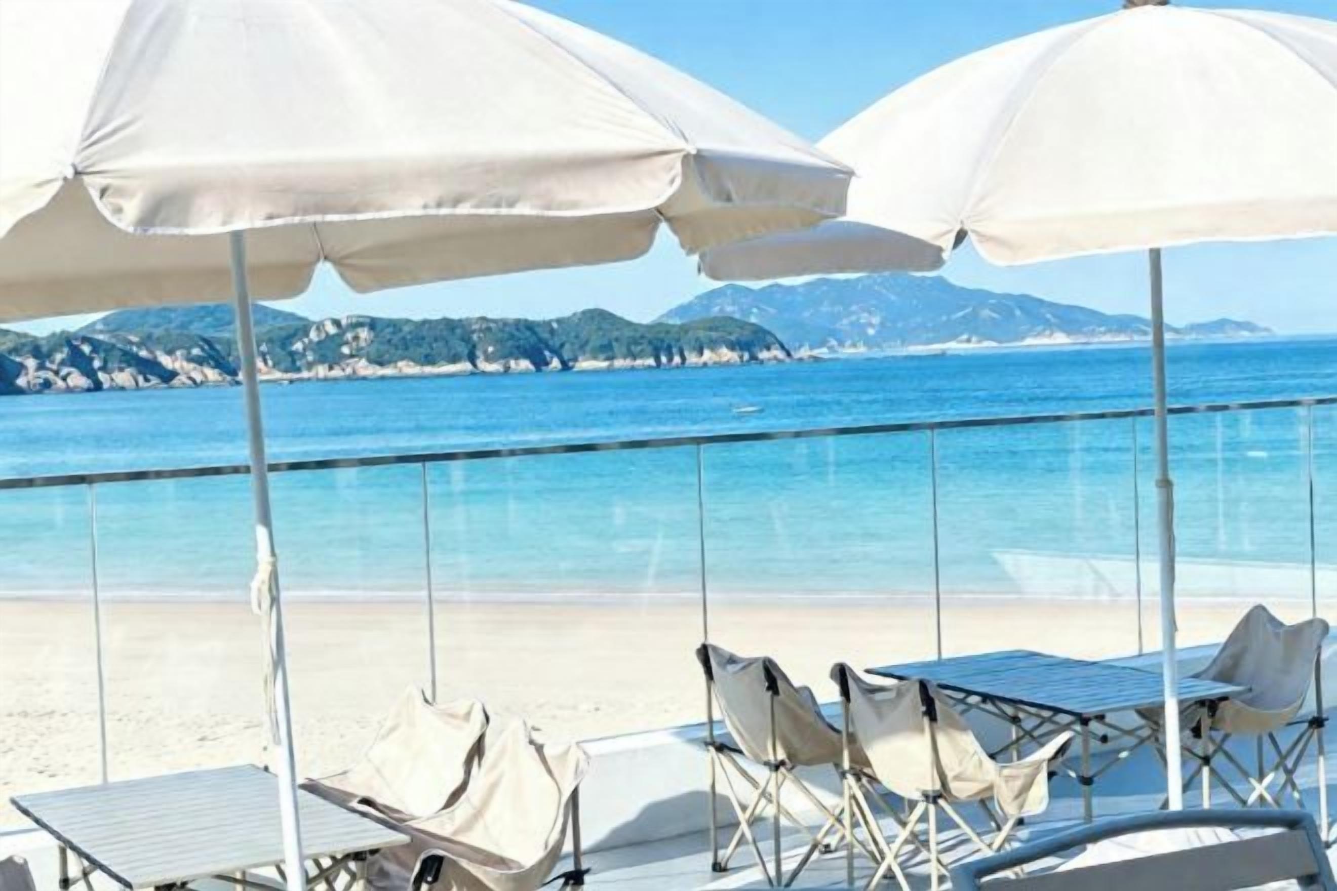 A beachside seating area with umbrellas and chairs, overlooking a clear blue sea and distant mountains.
