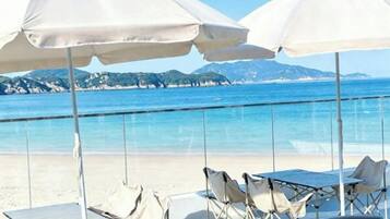 A beachside seating area with umbrellas and chairs, overlooking a clear blue sea and distant mountains.