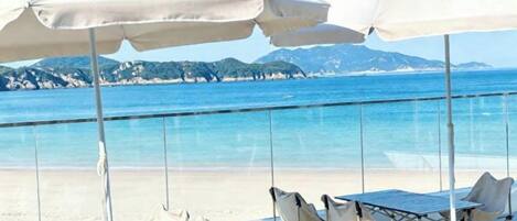 A beachside seating area with umbrellas and chairs, overlooking a clear blue sea and distant mountains.