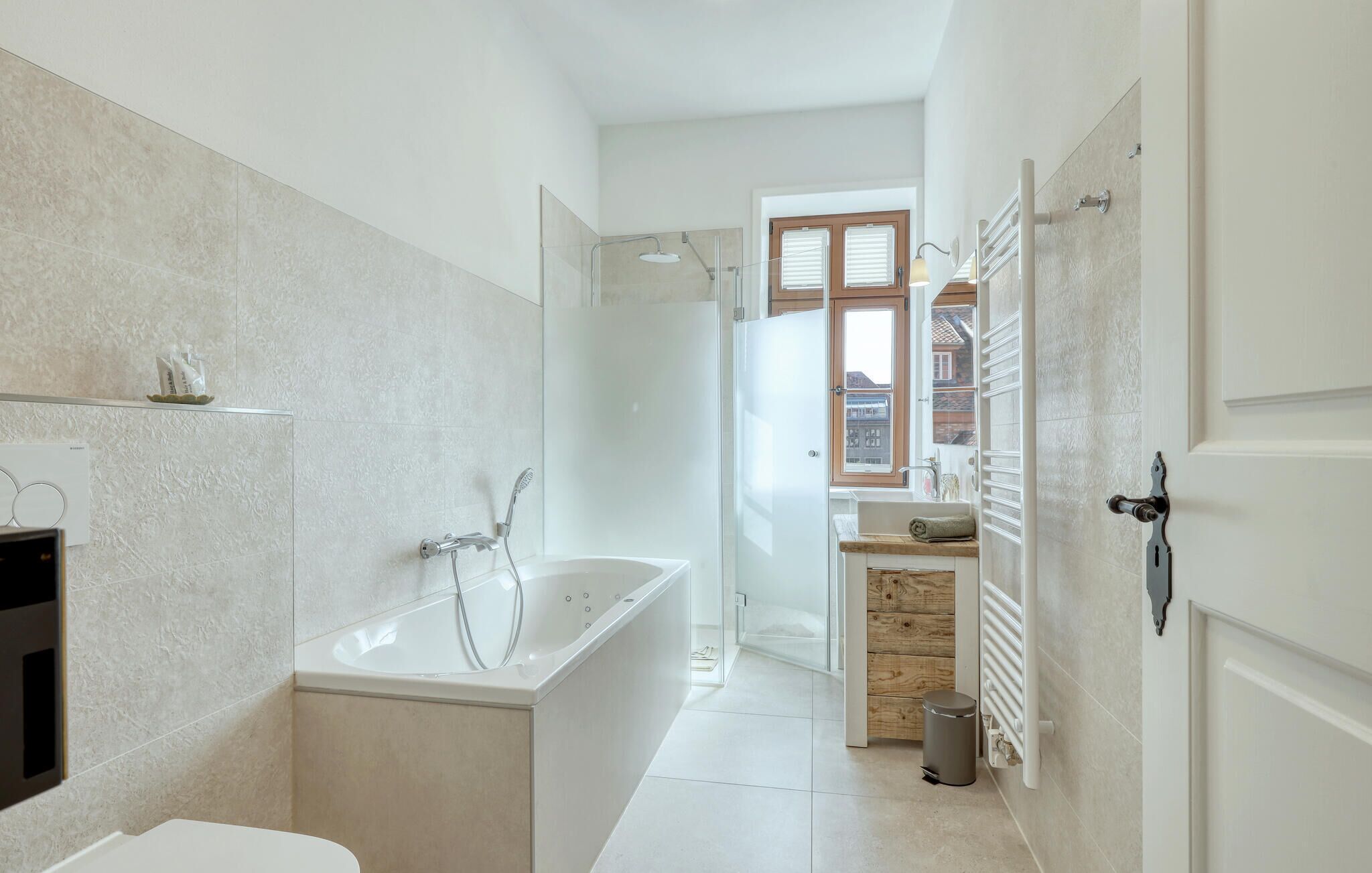 Une salle de bain moderne équipée d’une baignoire, d’une douche et de toilettes. On y trouve une fenêtre avec des stores, un porte-serviettes et un radiateur.