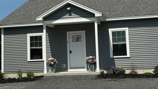 A single-story house with a gray siding exterior, a white front door, and two flower planters on either side of the entrance.