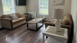 A living room with a brown sofa, a coffee table, and a lamp. There are two windows with blinds, a wall-mounted TV, and a radiator.