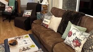A cozy living room with a brown sofa, a small table with a placemat, and a patterned rug. A staircase with wooden railings is visible in the background.