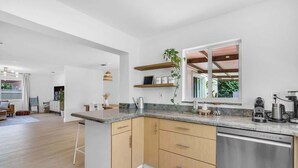 Una cocina moderna con armarios de madera, lavavajillas de acero inoxidable y encimera de granito. Hay estantes empotrados sobre la encimera y un ventanal que proporciona luz natural.