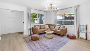 Una sala de estar moderna con un sofá seccional color beige, una mesa redonda y dos otomanas. Se observan ventanas amplias con cortinas, un lámpara en el techo y una puerta a la izquierda.