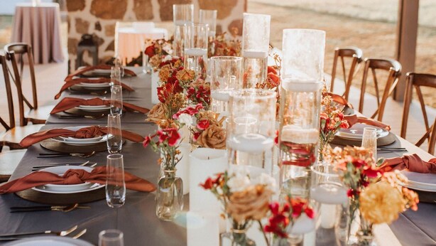 Una larga mesa de comedor lista para una comida, con copas transparentes, servilletas rojas y centros de mesa florales.