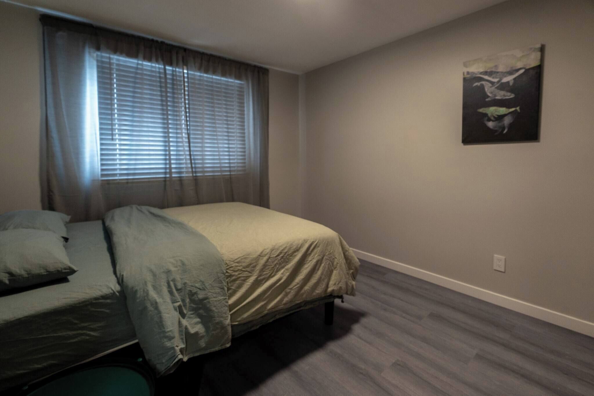Un dormitorio con cama, ventana con persianas, un cuadro en la pared y piso de madera.