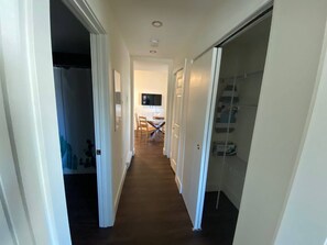 Um corredor estreito com piso de madeira, paredes brancas e uma porta que dá para um quarto com cama e televisão.