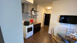 Una cocina moderna con estufa, microondas y lavamanos, un comedor con mesa de vidrio y sillas, y un televisor montado en la pared.