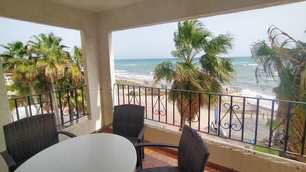 Un balcón con sillas de mimbre y una mesa, con vista a una playa con palmeras y el océano.