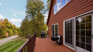 A wooden deck with a grill and a table, a sliding glass door, and a view of a road and trees. - Ski & Golf Spacious Log Cabin in Otsego Resort (Gaylord)