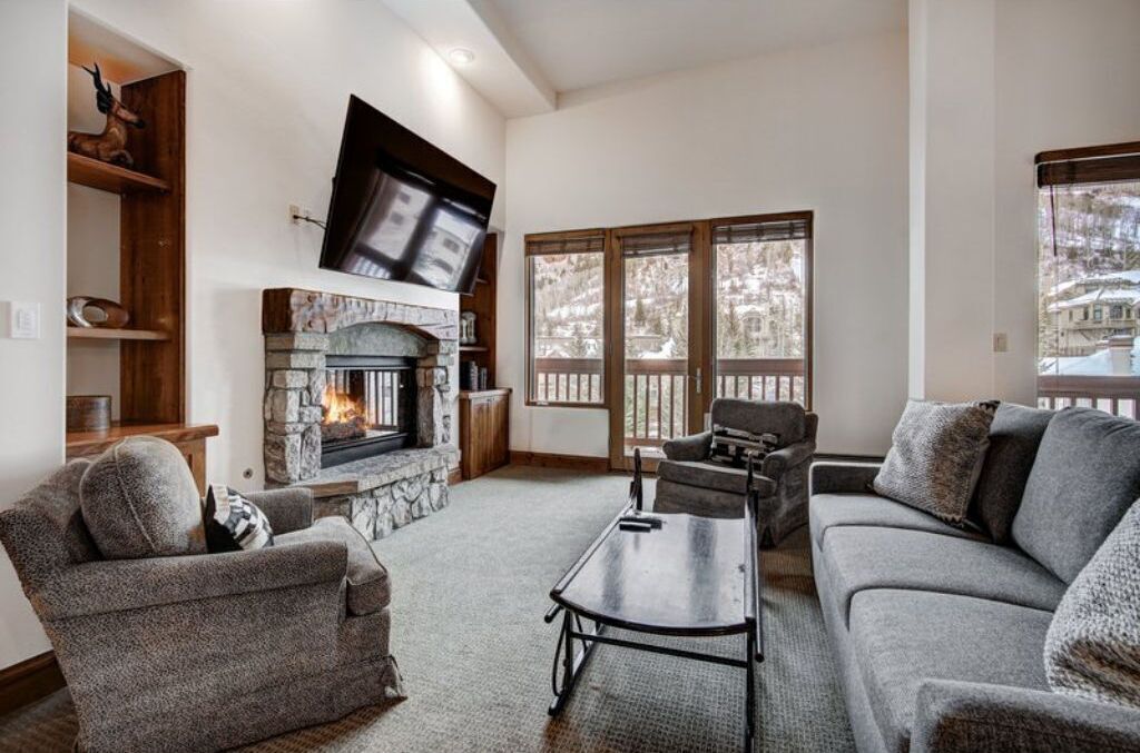 A cozy living room with a stone fireplace, a flat-screen TV mounted above it, and a large window with a view of a snowy landscape.