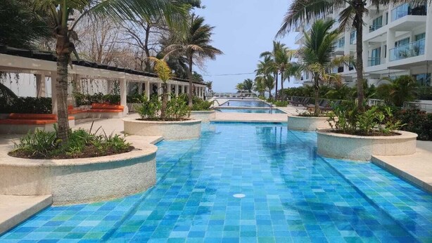 A swimming pool with a tiled floor, surrounded by palm trees and a building with balconies. - Apartamento en Cartagena de Indias, Edificio Morros 3 (Cartagena)