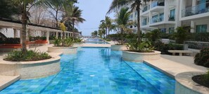 Ein Schwimmbecken mit Fliesenboden, umgeben von Palmen und einem Gebäude mit Balkonen.