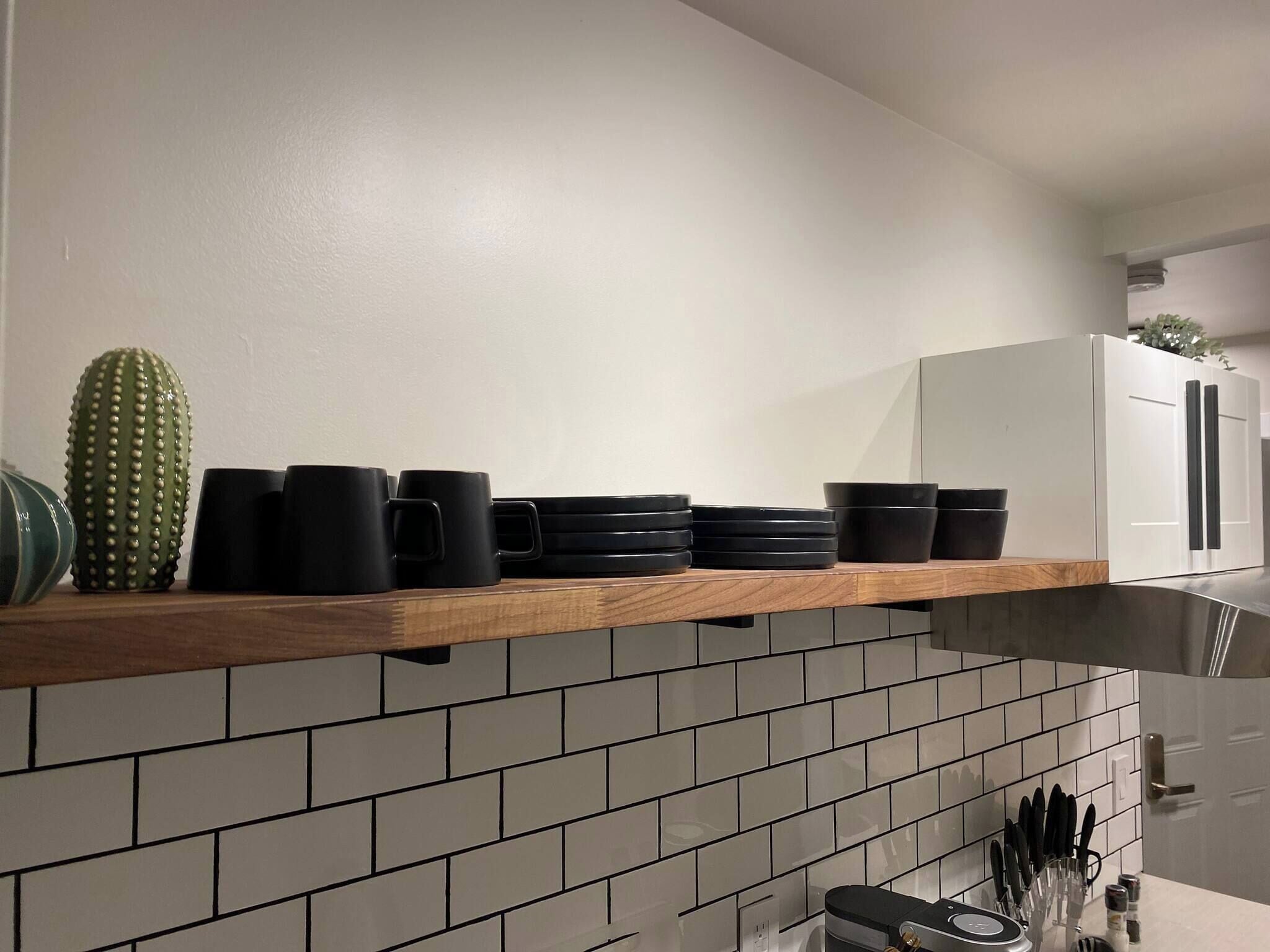A kitchen with a wooden shelf holding black mugs, a cactus, and a white refrigerator.